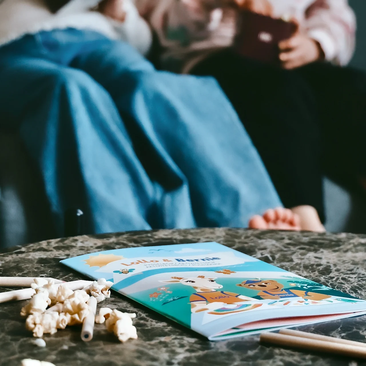 Coloring book with cartoon animals lies open on a marble table, surrounded by scattered popcorn and pencils. Two people sit on a sofa in the background.