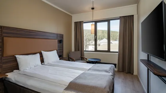A neatly made bed with a brown headboard, positioned in a hotel room. Natural light enters through a large window with curtains, beside a small seating area and a wall-mounted TV.