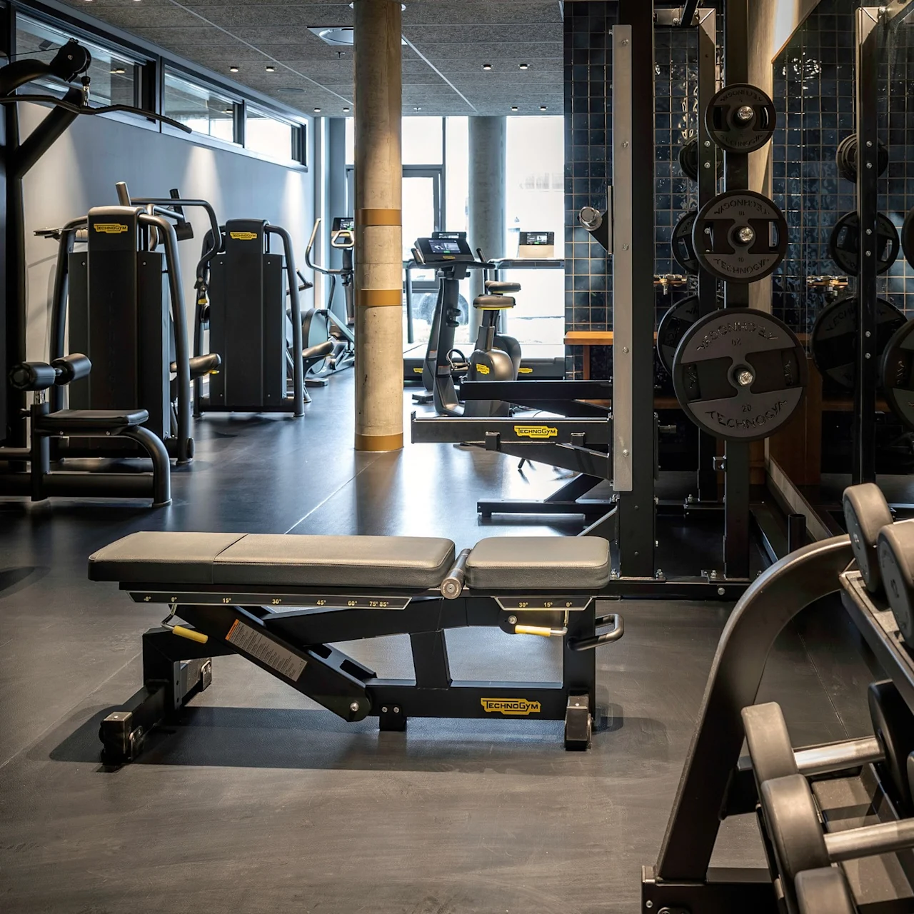 En vægtbænk står stille i et moderne fitnesscenter omgivet af forskellige træningsmaskiner og vægte, under lys naturlig belysning.