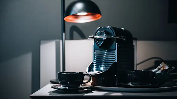 A coffee machine sits on a kitchen counter, next to two black cups on saucers under a sleek black lamp, creating a modern, cozy atmosphere.