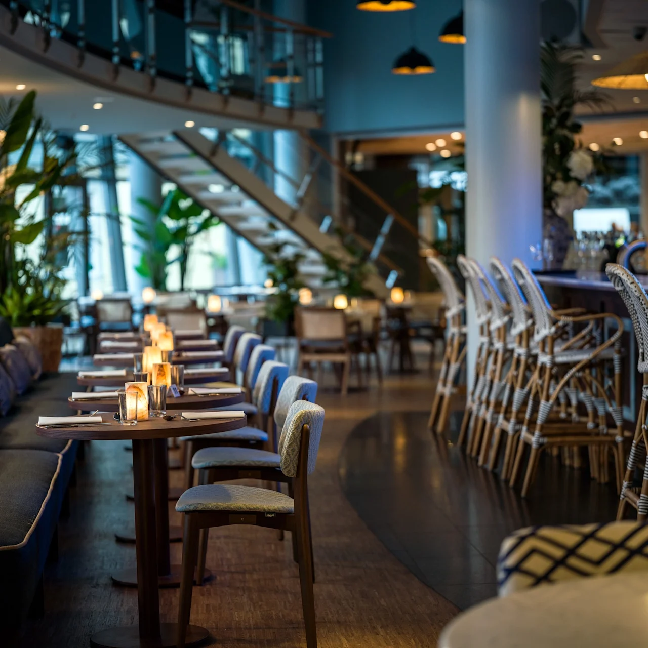 Borden är prydligt arrangerade med stolar och upplysta av ljus i en stilfull restaurang, med en svängd trappa och stämningsbelysning som skapar en mysig atmosfär.