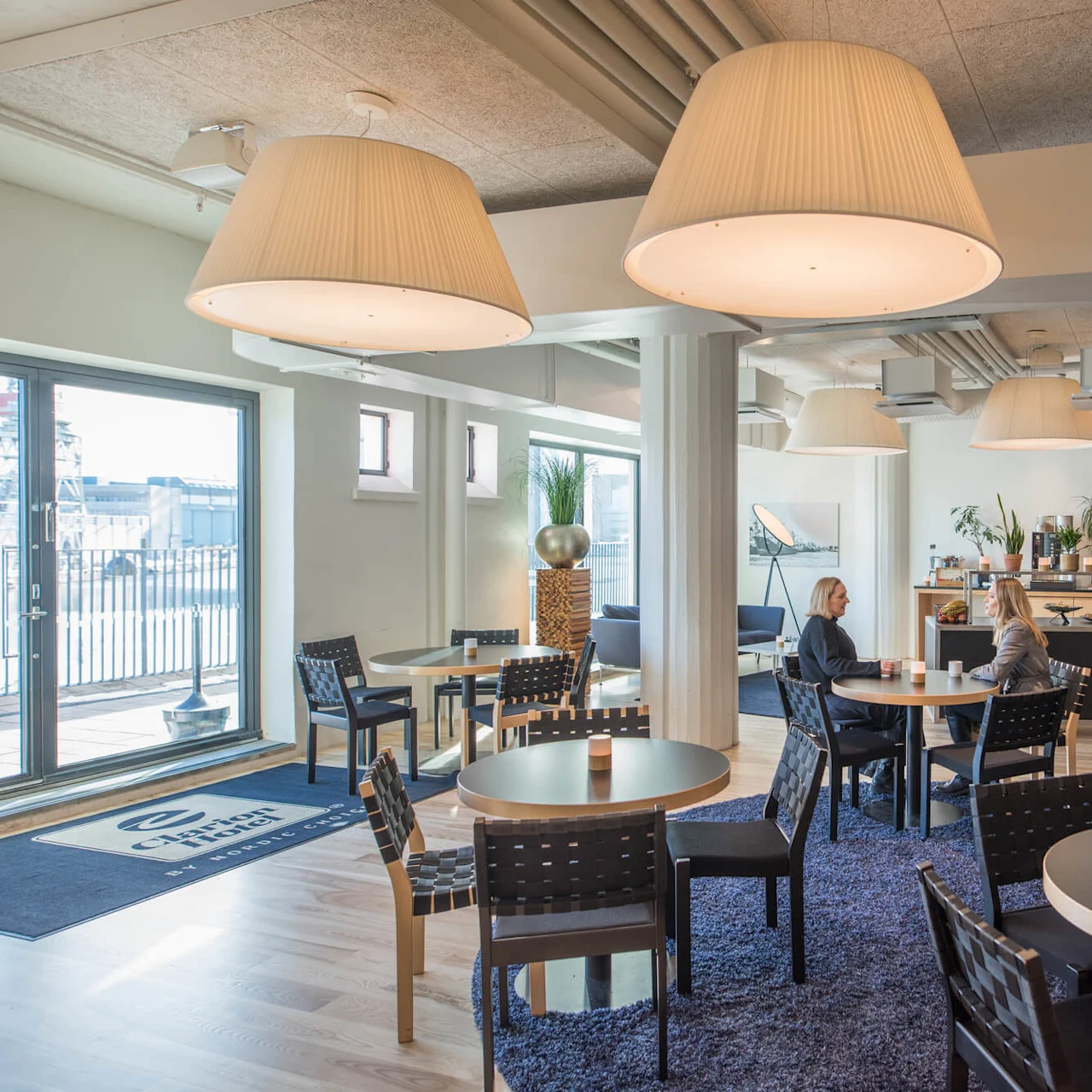 Et moderne, lyst caféområde med runde borde, flettede stole og store hængende lamper, har to personer der snakker. Glasdøre åbner til en balkon med industriel udsigt. Tekst på måtte: "Clarion Hotel".