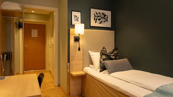 A cozy hotel room with a single bed, a desk, and a wardrobe area. The room features modern decor and warm lighting.