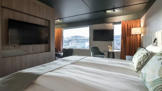 Siististi petattu sänky koristeellisilla tyynyillä on seinälle asennettujen televisioiden edessä modernissa hotellihuoneessa, josta on näkymä lumiseen, mäkiseen kaupunkimaisemaan suurten ikkunoiden kautta, oranssien verhojen kehystämänä.
