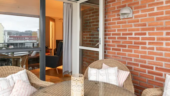 En koselig balkong med kurvstoler og et glassbord, med utsikt over byen og et innbydende innendørsområde.