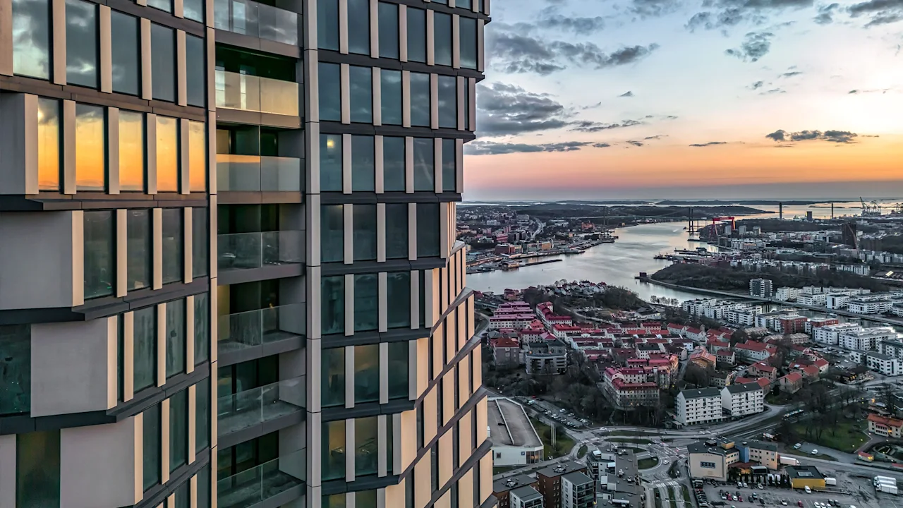 A building with glass windows reflects the sunset; adjacent is an urban landscape with red-roofed buildings, winding river, and sprawling cityscape under a cloudy sky.