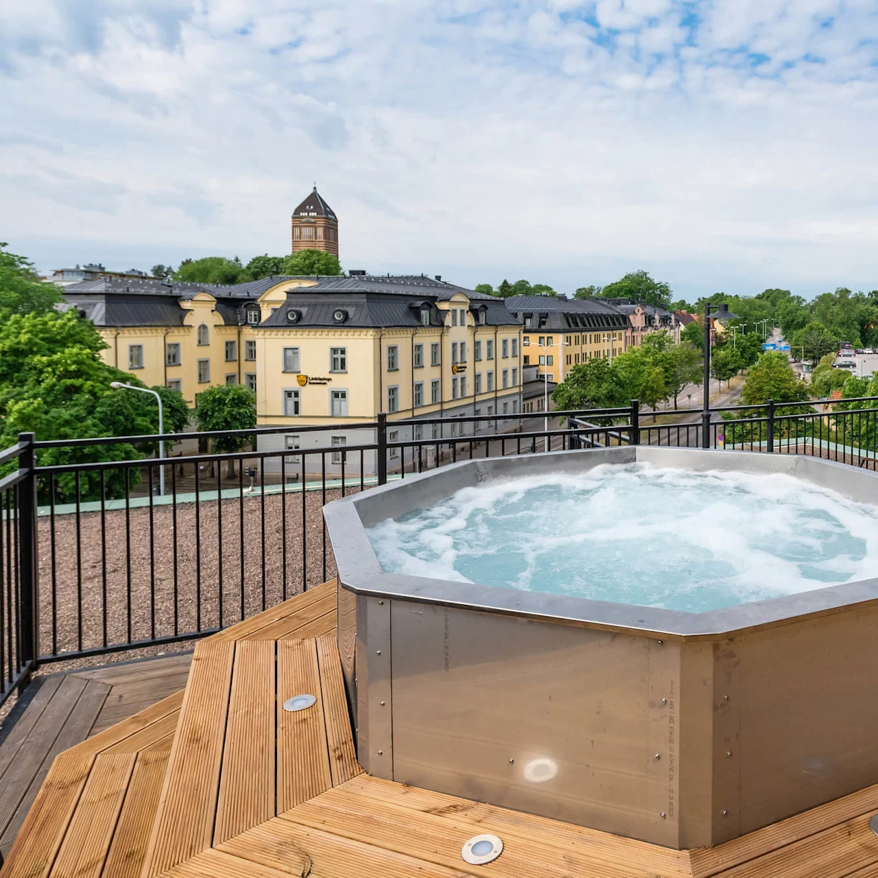 En bubbelpool med skummande vatten på en träterrass, med utsikt över en stadssilhuett med klassisk arkitektur och frodig grönska.
