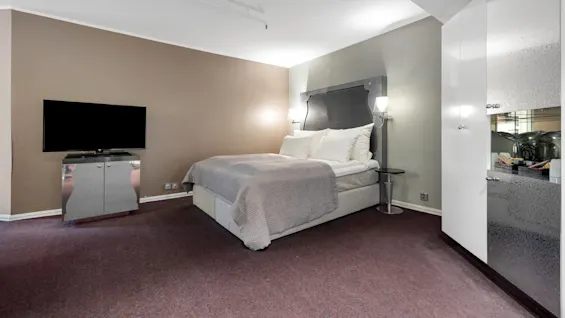 En seng med gråt sengetøj står i et moderne hotelværelse, flankeret af to væglamper; et fladskærmstv står på et skab modsat. Brun tæppe og hvide vægge omgiver.