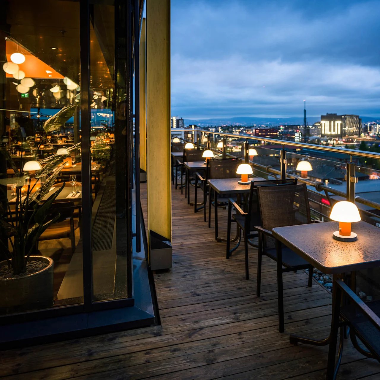 Outdoor dining tables with glowing lamps line a wooden terrace. The cityscape, visible at dusk beyond a glass railing, features a lit building displaying the word "TILIT."