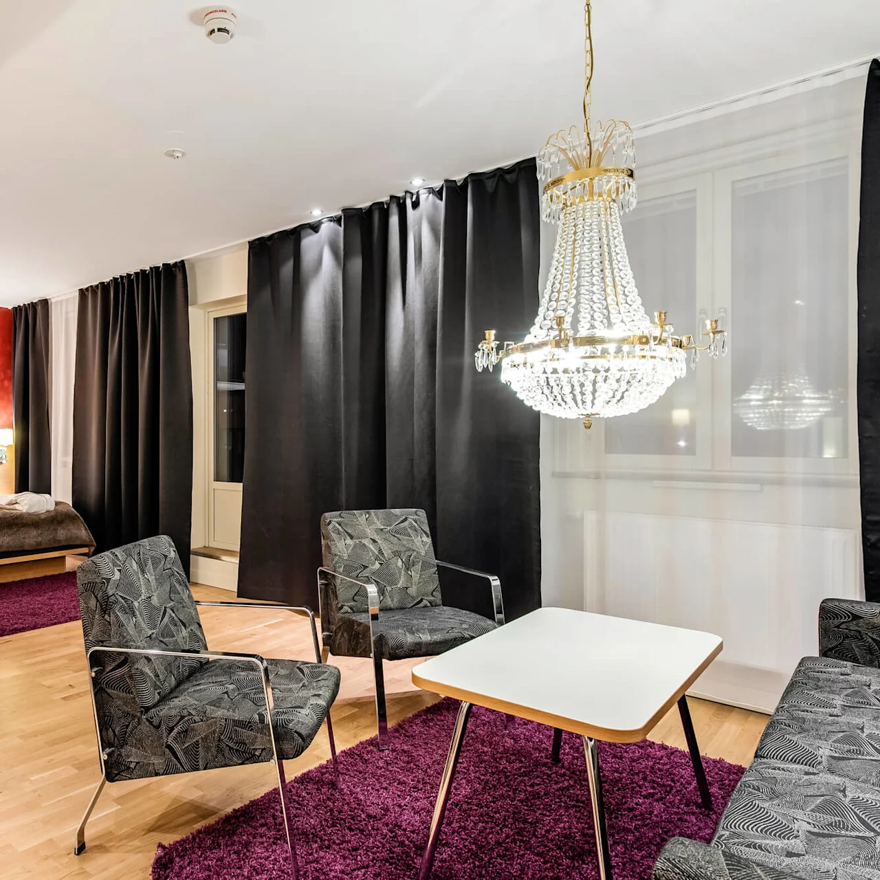 Et romslig hotellrom har en seng med et brunt teppe, en krystallkrone og et sitteområde med to stoler og et bord over et lilla teppe.