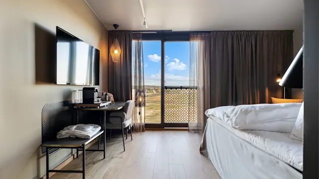 En pent seng står ved siden av gulv-til-tak-vinduer i et moderne hotellrom, med en TV på veggen, et skrivebord under og utsikt over et solfylt landskap.