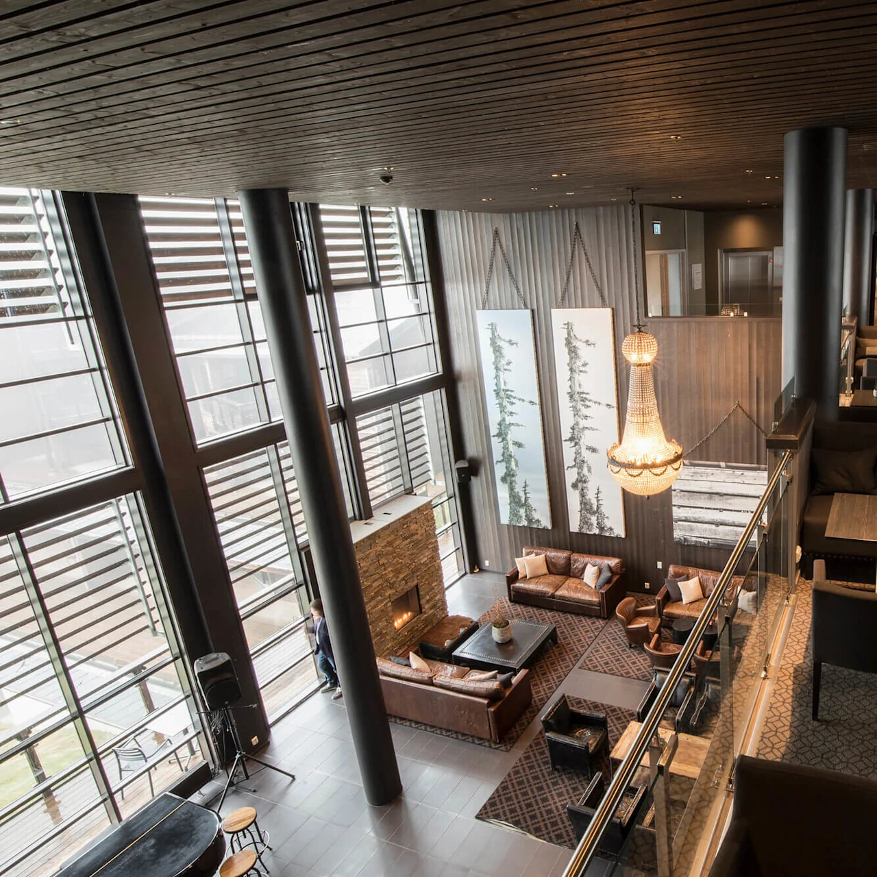 Elegant lounge area featuring leather sofas and a fireplace, illuminated by a large chandelier; surrounded by tall windows, art pieces, and tables with chairs, creating a cozy ambiance.