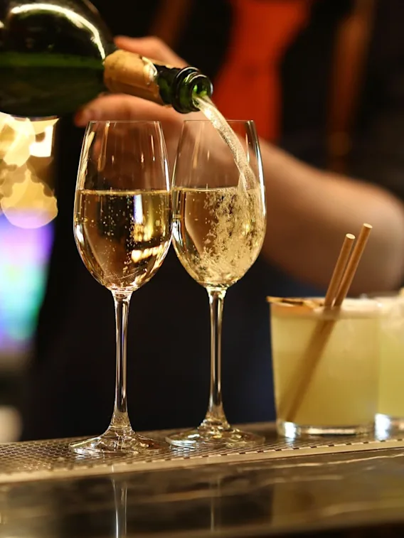 Champagne is being poured into two glasses on a bar, with a bartender in the background. Two cocktails with straws are next to the glasses, while warm lights create a cozy atmosphere.