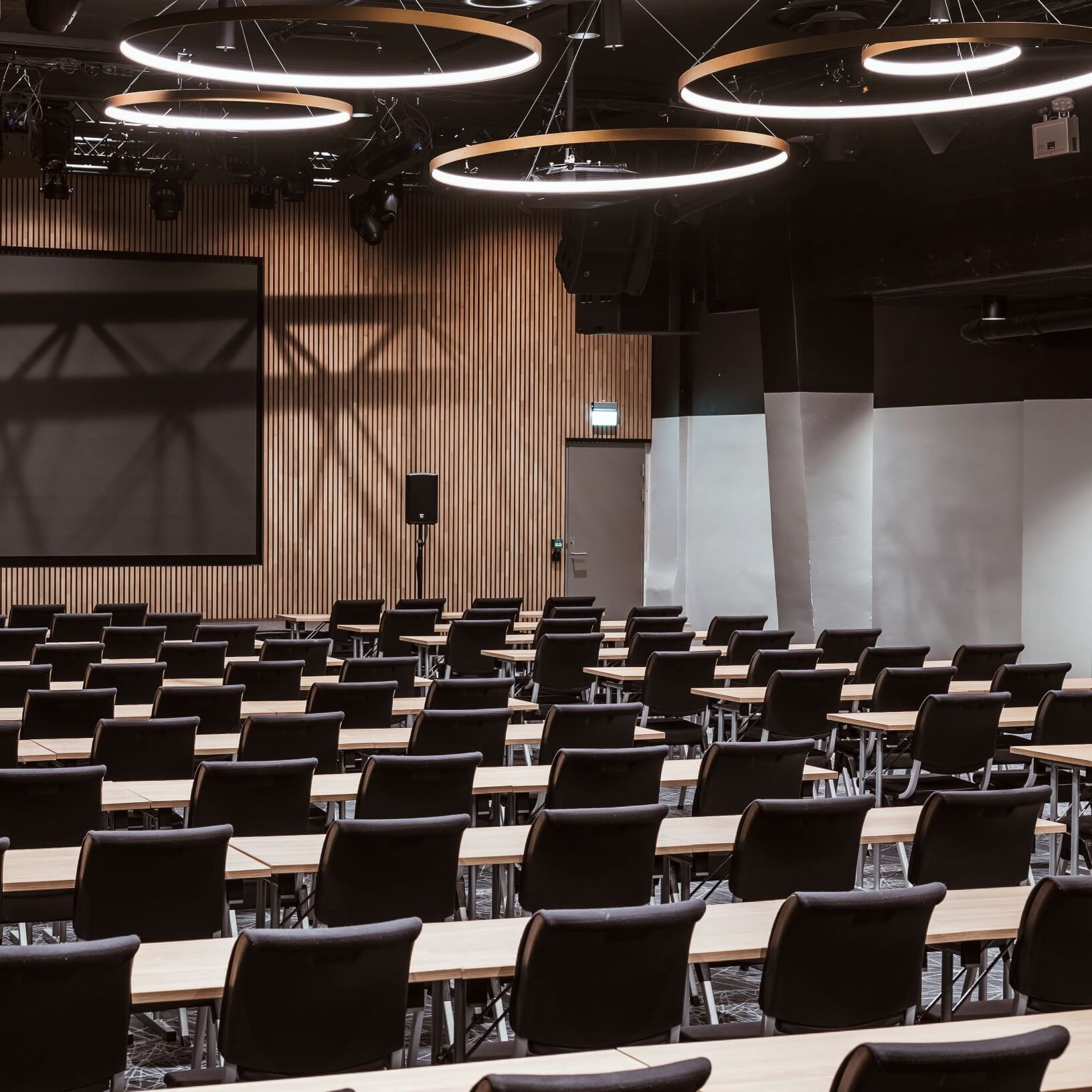 Rows of empty chairs facing a large screen in a modern conference room with wooden paneling and circular lighting fixtures.