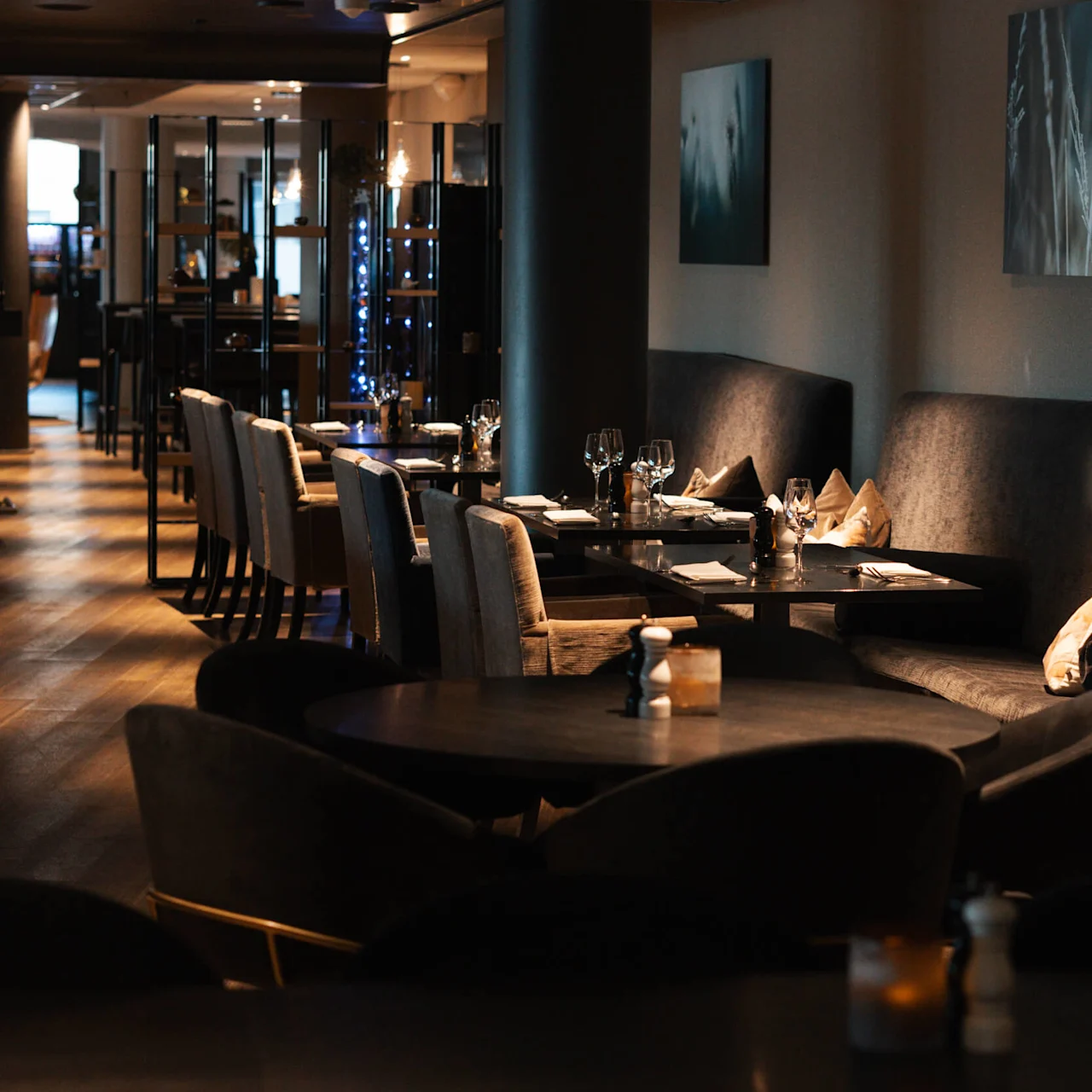 Elegante spisebord er dekket med glass og servietter, plassert i en dempet belyst moderne restaurant med polstrede seter og abstrakt kunst på veggene.