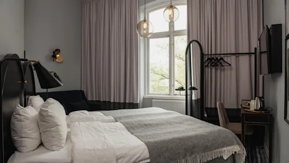 En prydligt bäddad säng med grått sängkläder står i ett modernt, stilrent sovrum. Gardiner täcker ett stort fönster; ett skrivbord, en stol och en klädhängare kompletterar den mysiga, minimalistiska inredningen.