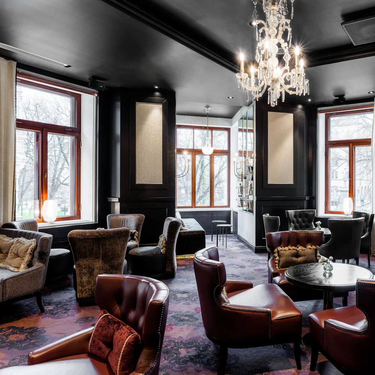 Luxurious lounge with leather chairs and round tables under a crystal chandelier, surrounded by large windows with curtains. Soft lighting and plush decor create an elegant atmosphere.