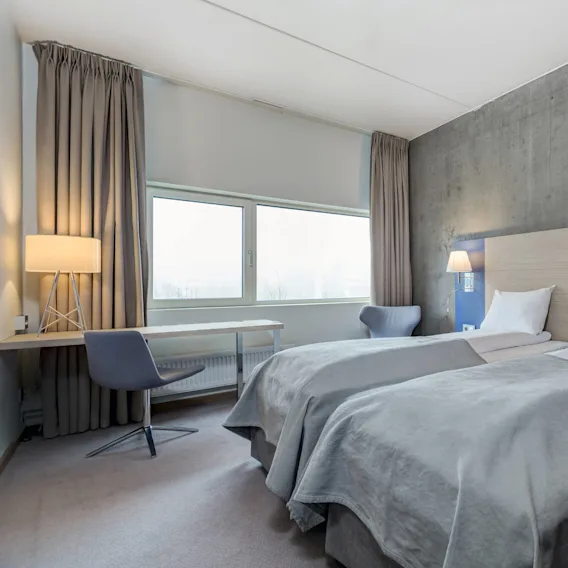 To pent oppredde enkeltsenger med hvitt sengetøy og grå tepper i et minimalistisk rom. En lampe og stol står ved siden av et vindu med lysegrå gardiner.
