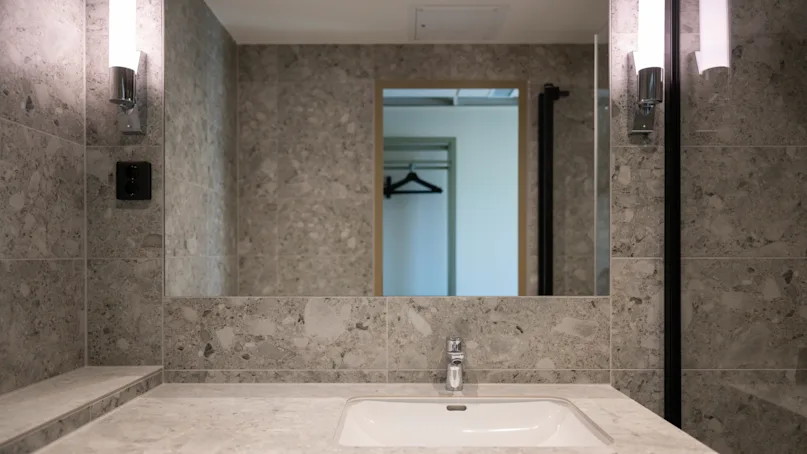 Sink sits under a large mirror, flanked by wall sconces, in a stone-tiled bathroom. A doorway in the mirror reflection reveals a closet with hangers.