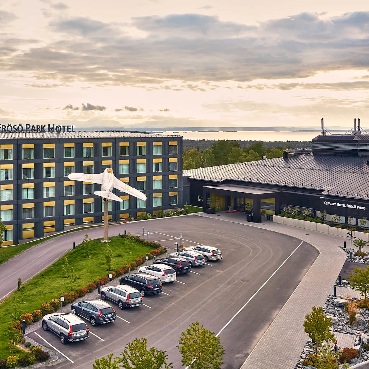 Ett hotell med skylten "Frösö Park Hotel" ligger omgiven av grönska, med en dekorativ flygplansmodell. Flera bilar är parkerade på parkeringen. Text innehåller "QUALITY HOTEL FRÖSÖ PARK."