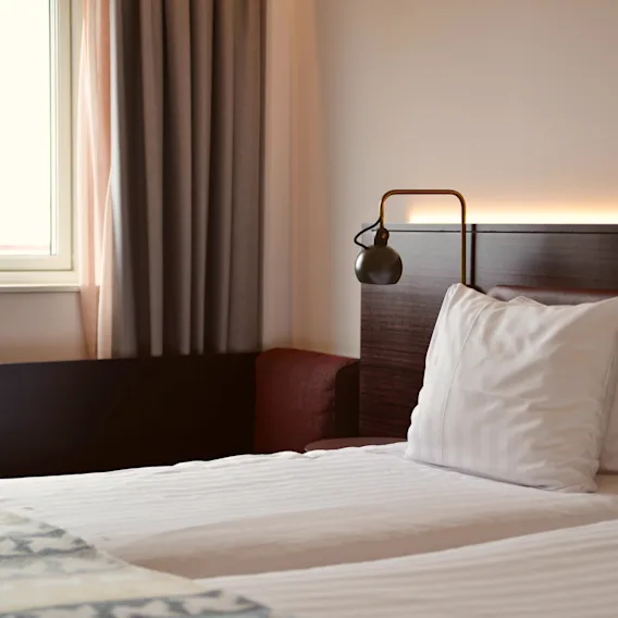 En säng med vita kuddar och en lampa på en trähuvudgavel i ett hotellrum bredvid ett fönster med gardiner.