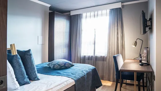 Et ryddig hotellrom med en pent oppredd seng, et skrivebord med en stol og et vindu som slipper inn naturlig lys.