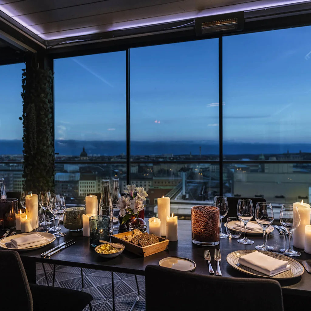 Ett ljusupplyst matbord med tallrikar, glas och snacks har utsikt över en stad genom stora fönster som visar en kvällshorisont.