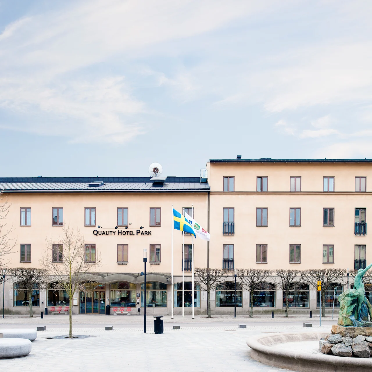 En lys brun bygning merket "Quality Hotel Park" står bak en brolagt plass. En statue på en steinplattform og to flaggstenger med svenske flagg står foran, blant løvløse trær.