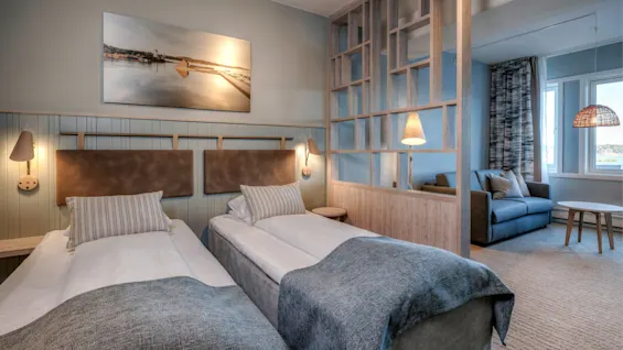 Two neatly made twin beds with gray blankets sit in a modern room, separated by a wooden partition from a sitting area with a sofa and round table.