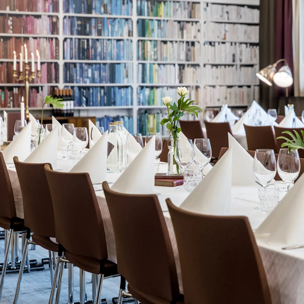 Elegantly set dining tables with white linens and napkins, surrounded by brown chairs, against a backdrop of bookshelves.