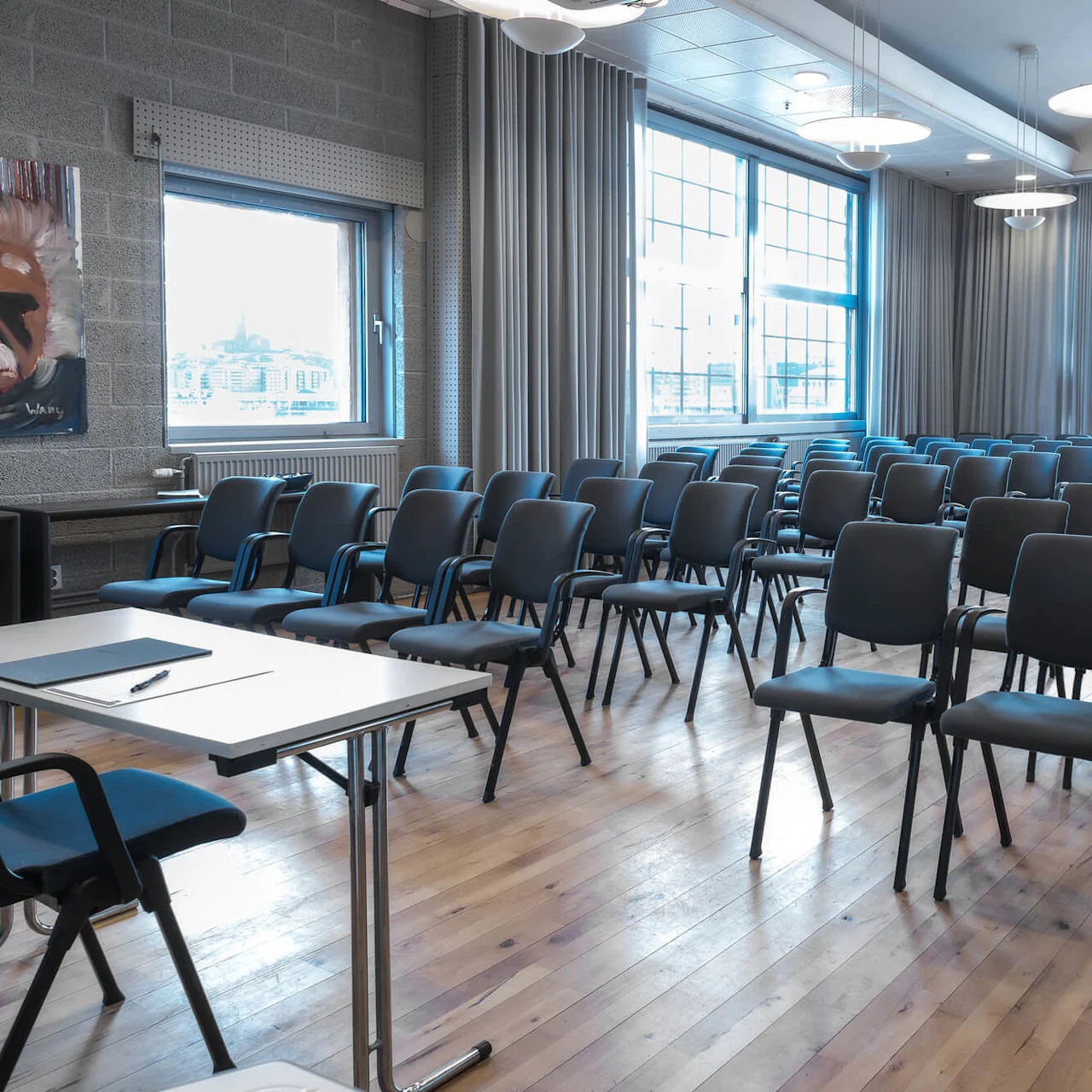Rows of empty black chairs face a blank presentation table with a notebook and pen, inside a well-lit conference room with large windows, wooden floor, and a painting on the wall.
