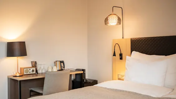 En snyggt bäddad säng med vita kuddar och beige filt bredvid ett skrivbord med en lampa, vattenkokare, koppar och en stol i ett mjukt belyst rum.
