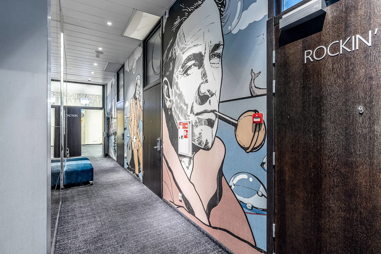 A hallway with a mural of a man smoking a pipe, surrounded by modern design elements. Text includes "ROCKIN'" on a door and "SATISFACTION" reflected on glass.