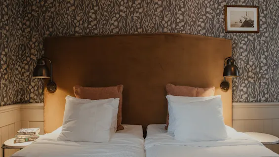 Viihtyisä hotellihuone kahdella sängyllä, lämpimällä sängynpäädyllä ja kuviollisella tapetilla, kutsuen rentouttavaan oleskeluun.