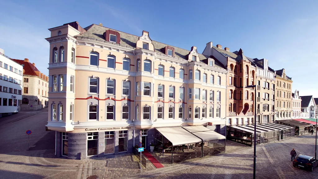 Elegant buildings line a sunlit, cobblestone square with awnings and a few pedestrians, in a quiet, historical town setting.