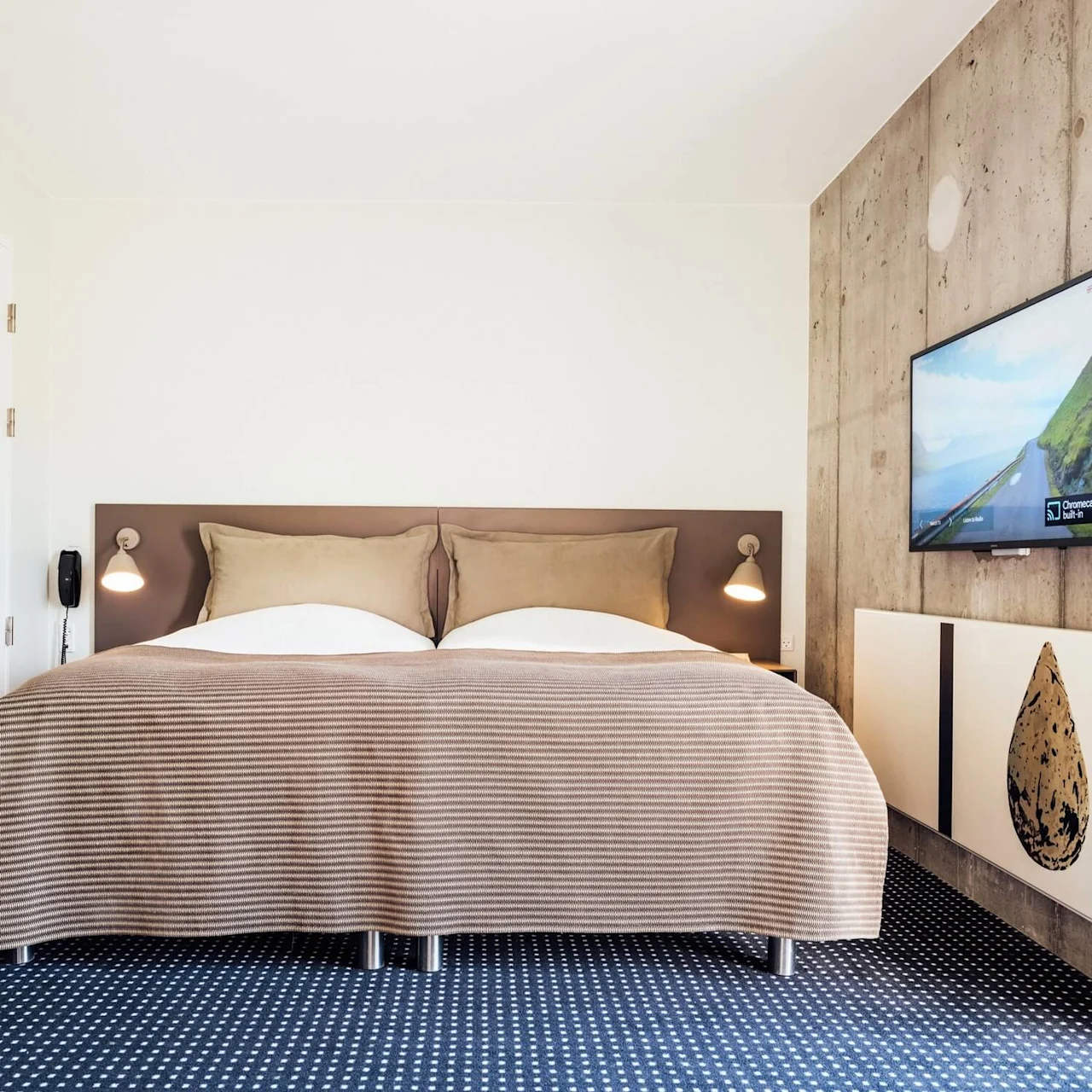 Seng under brunstribet tæppe, placeret mod en hvid og betonvæg med fladskærm, der viser landskab og "Chromecast Built-in." Moderne rum med væglamper og blåmønstret tæppe.
