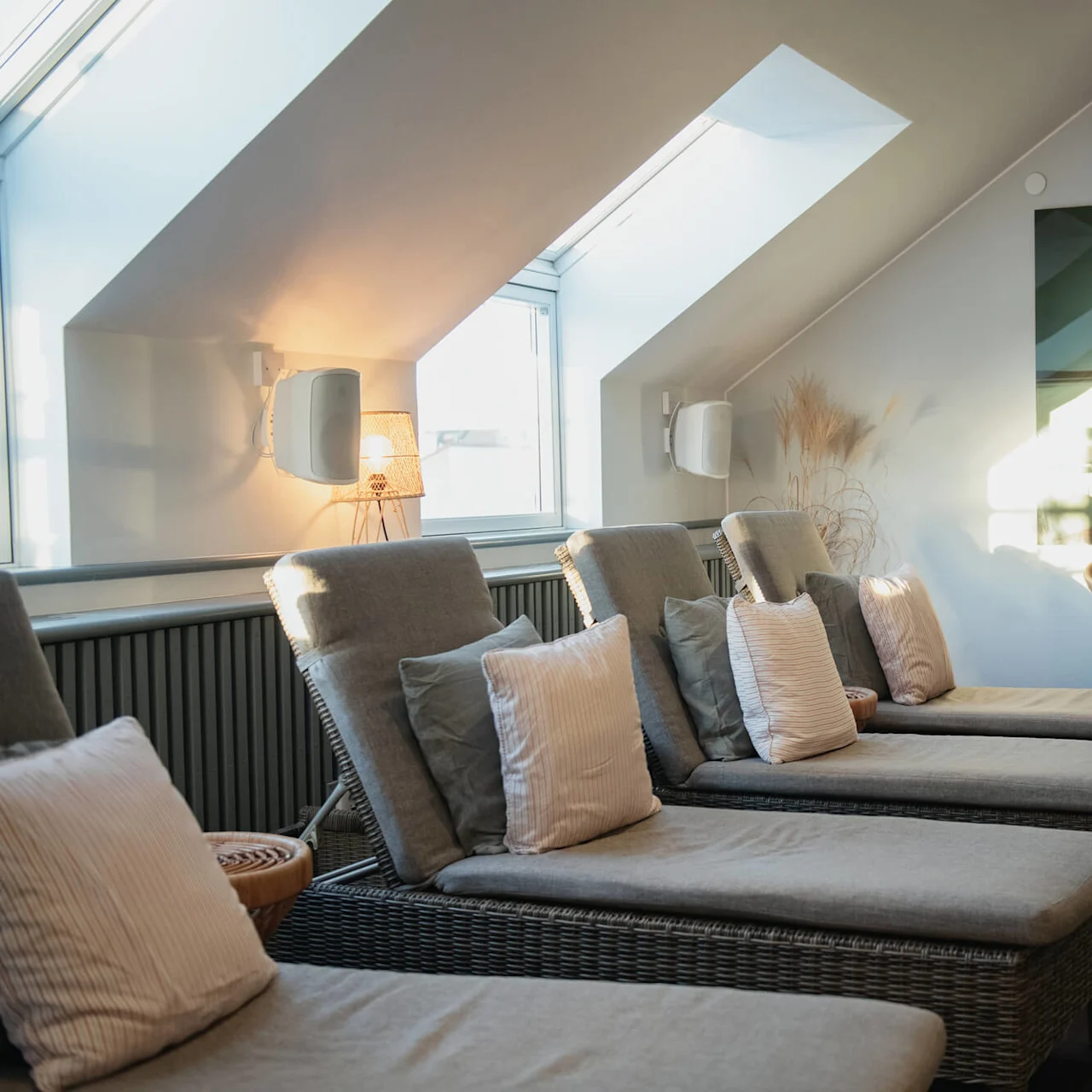 Lounge chairs with pillows rest under slanted skylights in a serene room with wooden floors, soft lighting, and a calming landscape painting on the wall.