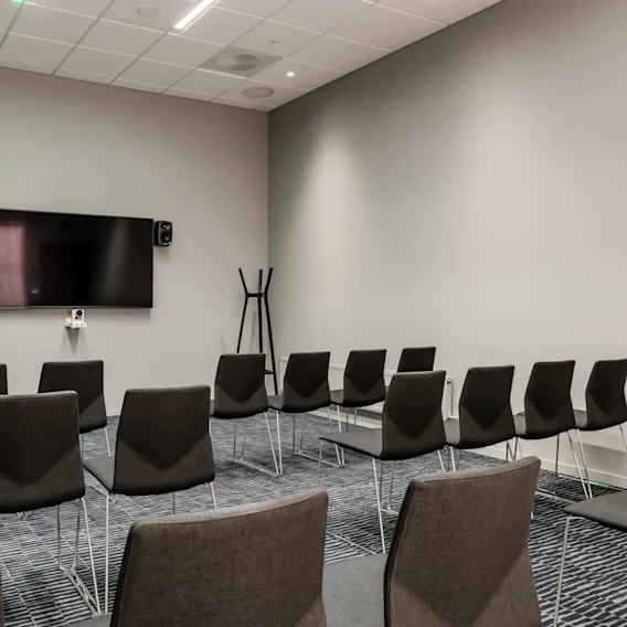 Chairs face a wall-mounted TV in a conference room, with speakers and a coat rack. The space is modern, featuring a blue-patterned carpet and white ceiling panels.