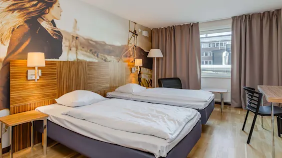 Kaksi erillistä sänkyä on siististi pedattu hotellihuoneessa. Suuri seinämaalaus naisesta, jonka hiukset hulmuavat, koristaa seinää. Ikkunan eteen on vedetty ruskeat verhot.
