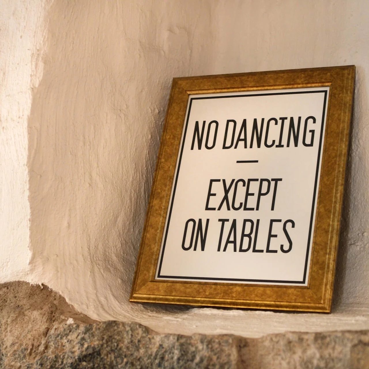 En inramad skylt på en strukturerad vägg med texten "NO DANCING - EXCEPT ON TABLES".