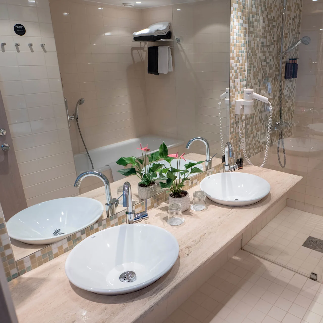 Två vita handfat står på en marmorbänk med en spegel ovanför, omgiven av beige kakelväggar. En liten krukväxt och toalettartiklar pryder utrymmet. En dusch och handdukar syns.