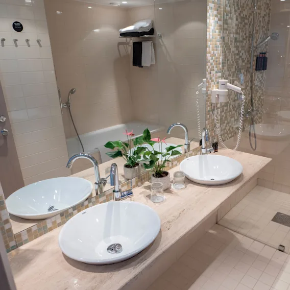 Två vita handfat står på en marmorbänk med en spegel ovanför, omgiven av beige kakelväggar. En liten krukväxt och toalettartiklar pryder utrymmet. En dusch och handdukar syns.