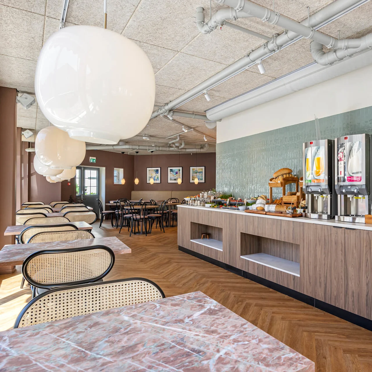 A modern cafeteria with self-service coffee machines, wooden tables and chairs, under large white globe lights.
