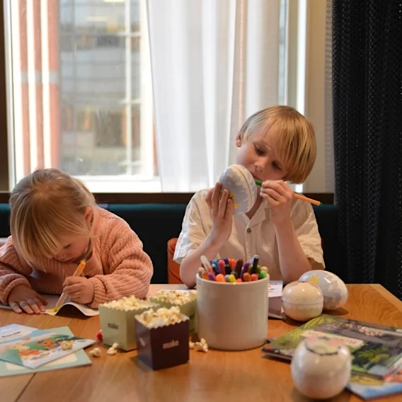 Two children engaged in drawing and coloring, seated at a wooden table cluttered with markers, books, and popcorn under a large window.
