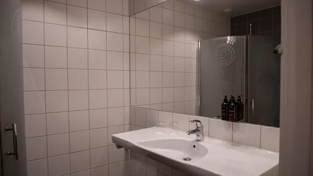 Handfat står orört under en spegelvägg i badrummet, omgivet av vita kakelplattor och upplyst av lampor ovanför; dusch och toalettartiklar syns i spegeln.