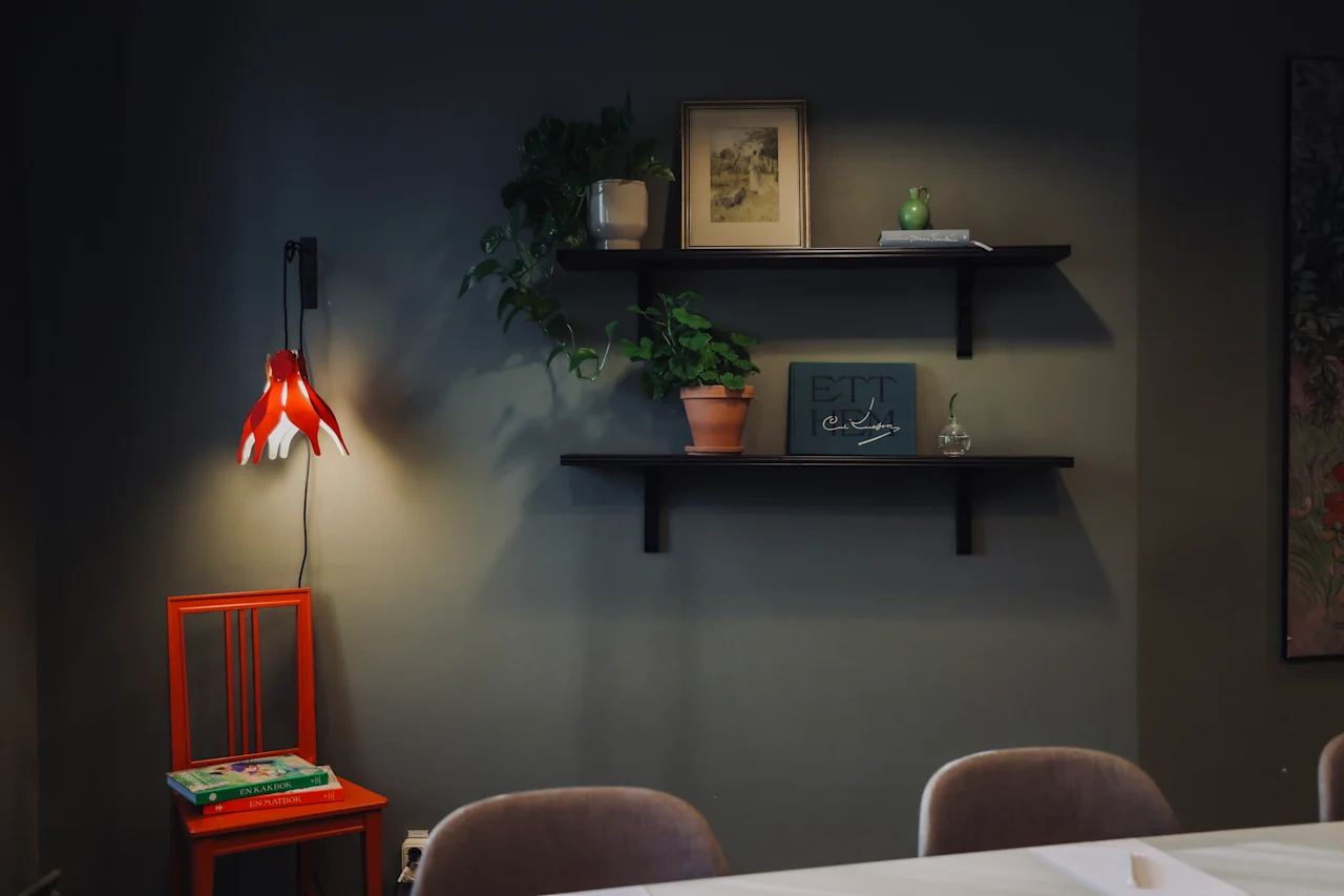 A wall with two shelves holds plants and framed art above a table. A red chair sits below, beside a red lamp, in a softly lit room.