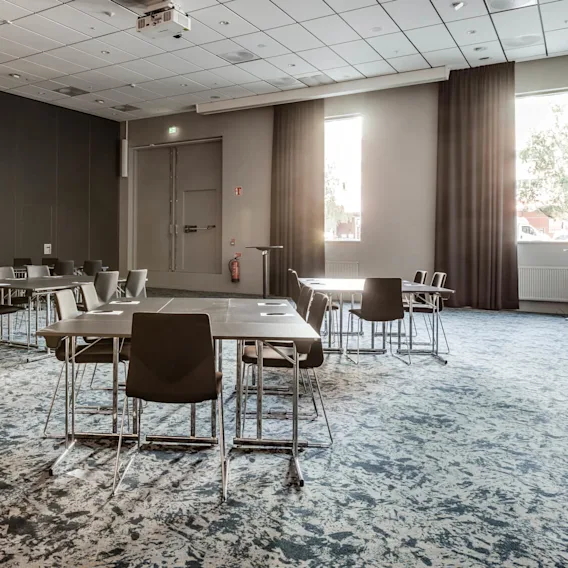 An arrangement of tables and chairs suggests a conference setup, in a room with patterned carpet and neutral walls. Sunlight streams through large windows, illuminating the space.