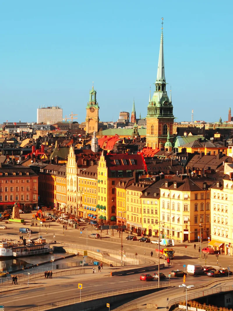 Colorful historical buildings line a bustling city square with people walking near a reflective waterway. Spires and towers rise against a clear blue sky, creating a picturesque urban scene.