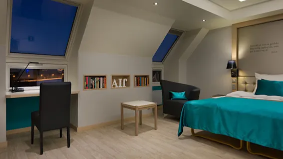 Et moderne soverom med en seng med tealfarger, en svart stol ved et skrivebord ved skråstilte vinduer og bokhyller. Varm belysning skaper en koselig atmosfære.