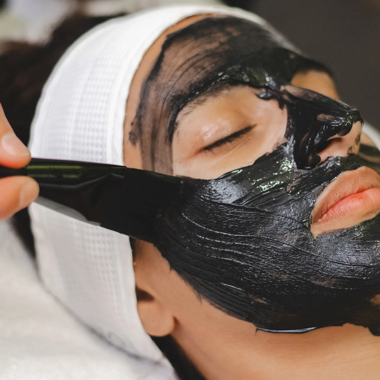 Person som får en ansiktsbehandling med svart leire, ligger med lukkede øyne, mens en hånd påfører masken med en børste i en spa-setting.